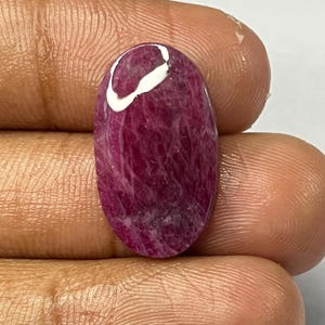 May include: An oval-shaped, polished gemstone with a deep magenta hue and a white, curved inclusion near the top. The stone has a smooth, glossy surface and is held in a hand against a plain white background. This gemstone is likely for jewelry making.