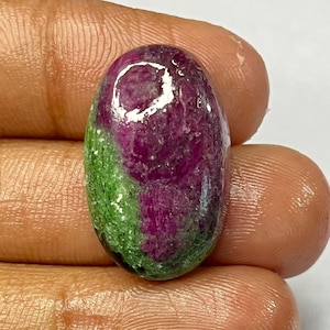 May include: An oval-shaped gemstone with a polished surface. The stone features a deep magenta hue with a section of green. The gemstone is held in a hand against a white background. The stone is likely a ruby.