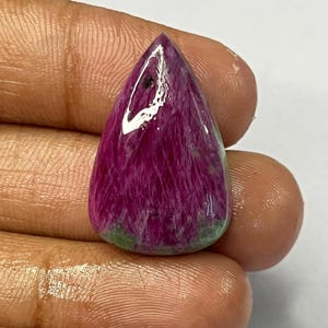 May include: A polished, teardrop-shaped gemstone with a deep magenta hue, veined with lighter pink and a touch of green at the base. The gem is held in a hand, showcasing its smooth, reflective surface. This gemstone is likely for jewelry making.