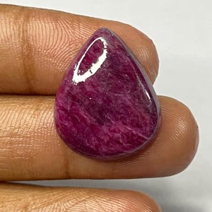 May include: A polished, teardrop-shaped gemstone in a deep magenta hue. The stone has a smooth, glossy surface with visible internal patterns. The gem is held between fingers, showcasing its size and shape. This could be a cabochon for jewelry making.