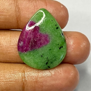 Puede incluir: Una gema en forma de lágrima con una llamativa combinación de colores. La piedra presenta una base verde vibrante, salpicada de inclusiones negras, y un prominente centro magenta. Esta piedra pulida es ideal para la creación de joyas.