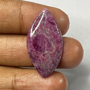 May include: A polished, marquise-shaped gemstone with a deep magenta hue. The stone has a glossy surface and a textured interior, showcasing natural patterns. The gemstone is held in a hand, highlighting its size and shape.