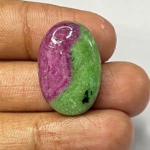May include: An oval-shaped gemstone, split into two sections. One side is a deep magenta, the other a vibrant green. The stone has a polished surface and a smooth texture. A small black inclusion is visible on the green side.