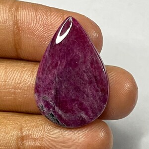 May include: A teardrop-shaped, polished gemstone in shades of deep red and magenta. The stone has a smooth, glossy surface with internal marbling and a small, dark inclusion near the base. The gem is held in a hand against a neutral background.