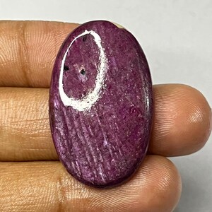 Puede incluir: Una piedra preciosa pulida, de forma ovalada, con un tono magenta intenso. La piedra tiene una superficie texturizada con estrías visibles y una pequeña inclusión oscura. La piedra preciosa se sostiene en una mano sobre un fondo blanco.