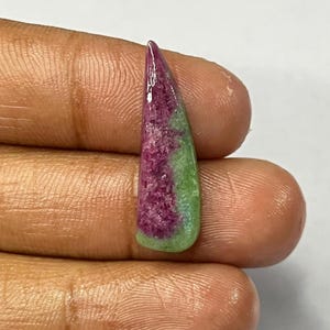 May include: A polished gemstone with a pointed top and a rounded base. The stone features a gradient of colors, including deep red, light green, and a touch of purple. The gemstone is held in a hand against a white background.