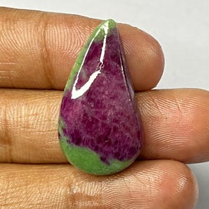 May include: A polished gemstone with a teardrop shape. The stone features a vibrant mix of colors, including deep magenta, green, and white. The magenta area has a textured appearance, while the green section is smooth. The gemstone is held in a hand.