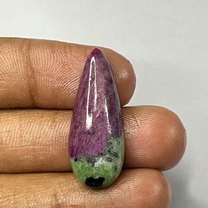 May include: A polished gemstone with a teardrop shape. The top portion is a deep magenta color, transitioning to green and black at the base. The stone is held in a hand against a white background.