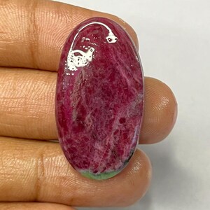 May include: An oval-shaped gemstone with a deep red hue, featuring a marbled texture and a hint of green at the base. The polished surface reflects light, showcasing the stone's natural patterns. This gemstone is a unique piece for jewelry making.