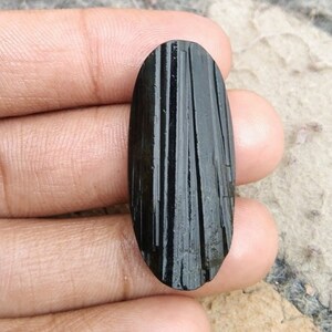 May include: A black, elongated gemstone with vertical striations. The polished stone is held in a hand, showcasing its dark color and textured surface. The gemstone's shape is oval, and it appears to be a natural mineral specimen.