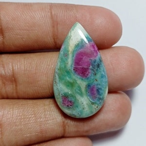 May include: A teardrop-shaped gemstone with a polished surface. The stone features a mix of colors, including green, blue, and white, with vibrant red spots. The gemstone is held in a hand against a white background.