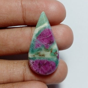May include: A teardrop-shaped gemstone with a mix of colors. The top is a light green, transitioning to white and then a vibrant magenta at the base. The stone is held in a hand.