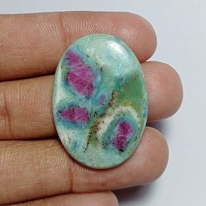 May include: Oval-shaped gemstone with a light green base and vibrant pink and red inclusions. The stone has a polished surface and is held in a hand, showcasing its natural beauty. The gemstone's unique color pattern makes it a striking piece.
