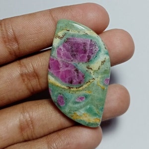 May include: A polished gemstone with a unique pattern. The stone features a mix of colors, including a light green base with vibrant pink and red inclusions. The stone is held in a hand, showcasing its natural beauty and intricate details.