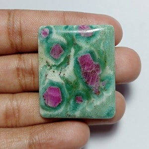 Peut inclure: Une pierre précieuse carrée avec une base vert clair, présentant des inclusions roses et rouges vibrantes. La pierre a une surface polie et un motif unique et organique. La pierre est tenue dans une main.
