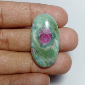 May include: Oval-shaped gemstone with a light green base and a prominent pinkish-red center. The stone has a polished surface, showcasing a swirling pattern of colors. The gemstone is held in a hand, highlighting its size and shape.