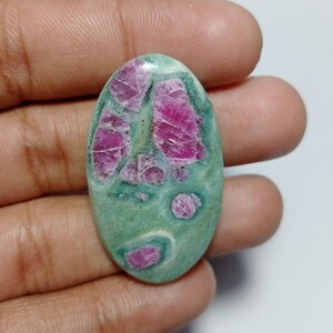Peut inclure: Pierre précieuse ovale avec une base vert clair et des inclusions magenta vives. La surface de la pierre présente un aspect moucheté, les zones magenta ressemblant à des structures cristallines. La pierre est tenue dans une main, mettant en valeur sa taille et ses variations de couleur.