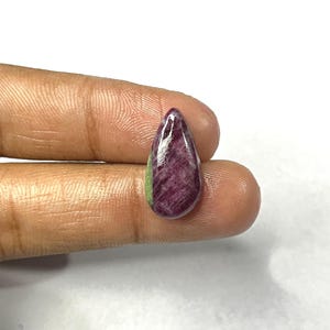 Puede incluir: Una gema pulida con forma de lágrima. La piedra presenta un color burdeos intenso con una franja verde en la base. La gema se sostiene entre dos dedos sobre un fondo blanco.