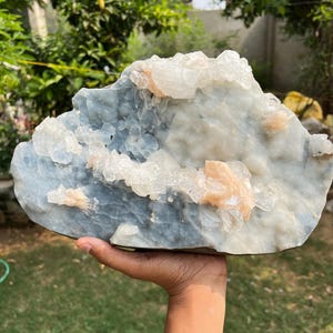 May include: A large, irregularly shaped crystal formation with a light blue base and clusters of white and peach-colored crystals. The natural stone is held in a hand against a backdrop of green foliage.