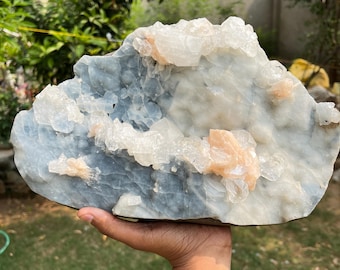 May include: A large, irregularly shaped crystal formation with a light blue base and clusters of white and peach-colored crystals. The natural stone is held in a hand against a backdrop of green foliage.