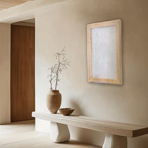 Op de afbeelding: Rechthoekige wandkunst met een licht geweven textuur en een natuurlijke houten lijst. Het kunstwerk hangt aan een beige muur. Een bank met stenen steunen en een vaas met een plant staan op de voorgrond.