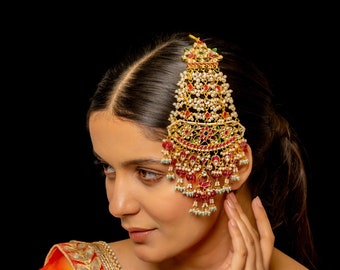May include: A large, ornate gold hair accessory featuring intricate filigree work, embedded with small white pearls, vibrant red stones, and green accents. The piece has a tiered, fan-like structure with dangling beads and teardrop-shaped pendants.