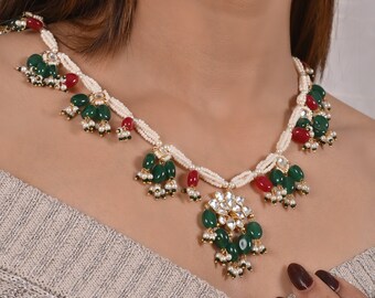 May include: A multi-strand necklace featuring small white seed beads, accented with dangling clusters of oval green and red beads. The center pendant showcases a floral-shaped arrangement of clear stones surrounded by green and red beads.