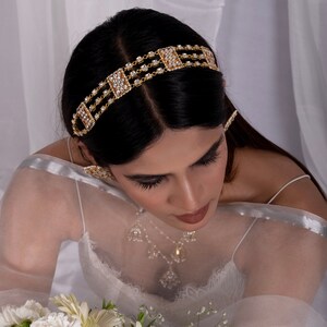 May include: A gold-toned headband featuring three parallel rows of small, clear, sparkling stones and rectangular, stone-encrusted accents. The piece is worn across the top of the head, showcasing a metallic finish and a repeating geometric pattern.