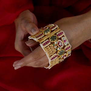 May include: A wide gold-toned hinged cuff bracelet featuring rectangular red stones, circular green stones, and intricate filigree metalwork. The edges of the cuff are lined with small white seed pearls, creating a detailed and ornate decorative accessory.
