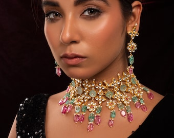 May include: A gold-toned choker necklace and matching drop earrings featuring floral motifs, inset with clear stones, and accented with dangling oval-shaped pink and green gemstones. The jewelry set displays intricate metalwork and a layered design.