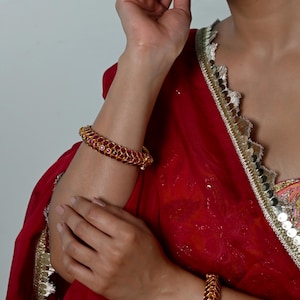 May include: A pair of gold-toned bangles featuring a repeating leaf-like pattern set with small, round, vibrant pink stones. The bangles are worn on the wrists, complementing a deep red fabric garment with a decorative metallic silver trim.