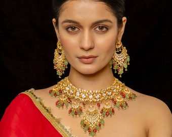 May include: A gold-toned necklace and matching drop earrings set featuring intricate metalwork, clear stones, and dangling light green and deep red beads. The necklace has a wide, ornate collar design with a central pendant and multiple hanging elements.