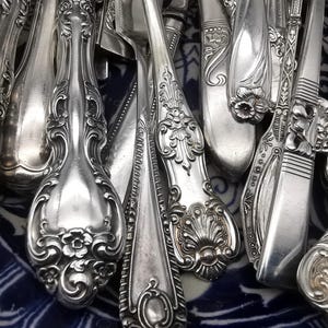 May include: A close-up of a collection of antique silver cutlery. The silverware features ornate floral and scroll designs, with a polished, reflective surface. The arrangement includes forks, spoons, and knives, showcasing various patterns and styles.
