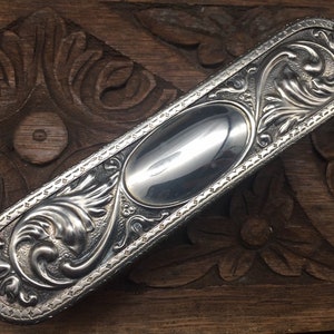 May include: A silver-plated, ornate, rectangular box with a decorative floral design and a smooth, oval-shaped interior.