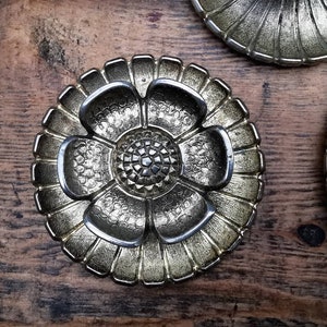 May include: A set of three gold-toned metal decorative trays with a flower design. The trays are round and have a raised edge. The flower design is in the centre of each tray and has five petals.