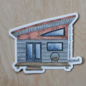 May include: A watercolor illustration of a small house with a metal roof, two windows, and a door. The house is on a wooden platform with a small fire pit in front. The text "DREAM SMALL" is written on the side of the house.