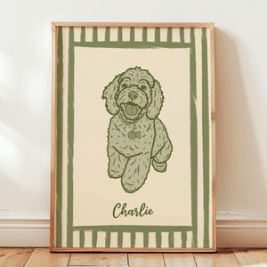 May include: A framed print featuring a smiling dog illustration in shades of green and beige. The dog has a collar with a tag and the name "Charlie" is written below the dog in a cursive font. The print is framed with a light brown wooden frame.