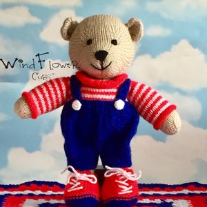 May include: A knitted teddy bear wearing red and white striped overalls and red shoes with white laces. The bear is sitting on a red, white, and blue crocheted blanket. The text "WindFlower Cubs" is visible in the background.