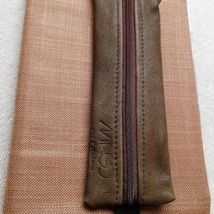 Könnte beinhalten: Ein braunes Leder-Etui mit dunkelbraunem Reißverschluss und einem schwarzen Gummiband ist an einem pfirsichfarbenen, stoffbezogenen Notizbuch befestigt. Auf dem Etui steht der Text "MCO aus Potsdam".