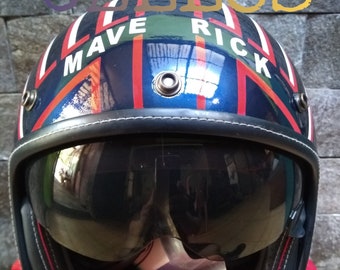 May include: A glossy blue motorcycle helmet featuring a dark tinted visor, a black trim, and a red and white striped pattern. The text MAVE RICK is printed in white across the top, while the word CELLOS appears in large letters at the top and bottom.