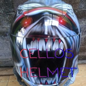 May include: A glossy black motorcycle helmet with a menacing, airbrushed design of a monster face. The design features sharp white teeth, red eyes, and the text "CELLOS HELMET" in colorful lettering. The helmet is displayed against a brick wall.