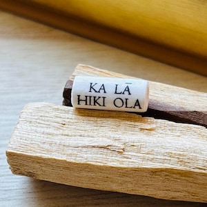 Puede incluir: Un objeto cilíndrico blanco con el texto "KA LĀ HIKI OLA" en letras negras descansa sobre un trozo de madera de color claro. La madera tiene una superficie texturizada. También es visible un trozo de madera marrón oscuro.