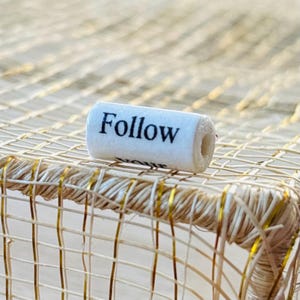 Pode incluir: Um objeto cilíndrico branco com a palavra "Follow" impressa em preto, repousando sobre uma superfície tecida. O objeto parece ser uma rolha ou material semelhante. O fundo apresenta uma textura tecida natural com detalhes dourados.