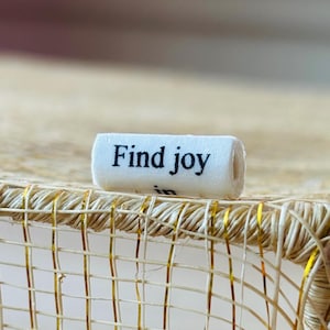 Peut inclure: Un petit rouleau de papier blanc avec les mots "Find joy" imprimés en noir. Le papier repose sur un panier tissé avec des accents dorés. Le panier est de couleur beige naturel.
