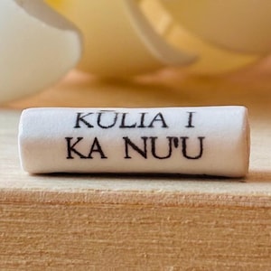 May include: A white, cylindrical object with the text "KULIA I KA NU'U" printed in black. The object is resting on a light brown surface, with a blurred background of a light yellow and white color.