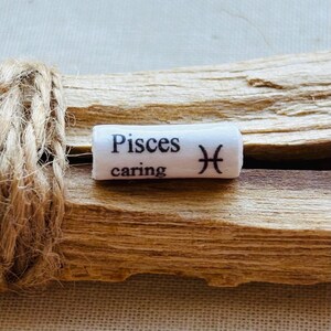 Könnte beinhalten: Ein weißer zylindrischer Anhänger mit den Worten "Pisces caring" und einem Fisch-Symbol in Schwarz. Der Anhänger ist auf einem Stück Treibholz befestigt und mit Bindfaden gebunden.