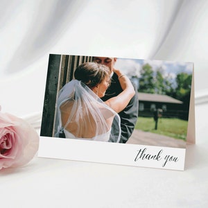 May include: A folded wedding thank you card with a photograph of a couple embracing. The card has a white border and the words "thank you" in elegant script. A pink rose is in the foreground.