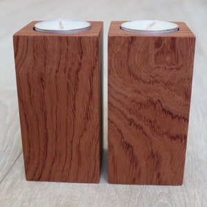 May include: Two wooden candle holders with a square design. Each holder is a reddish-brown color and has a circular indentation to hold a tea light candle. The wood grain is visible on the surface.