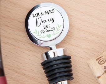 May include: A silver-toned metal bottle stopper featuring a circular top with a white insert. The insert displays the text MR & MRS Davies EST 20.08.23, accented by a green leaf illustration and a small heart. The stopper has a black ribbed rubber seal.