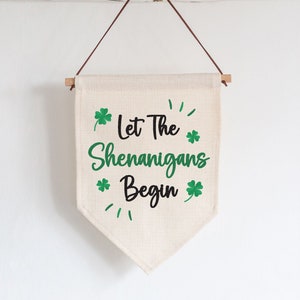 May include: A beige fabric wall hanging with the text "Let The Shenanigans Begin" in green and black, with green shamrock graphics. The banner is suspended by a wooden dowel and a brown leather cord.